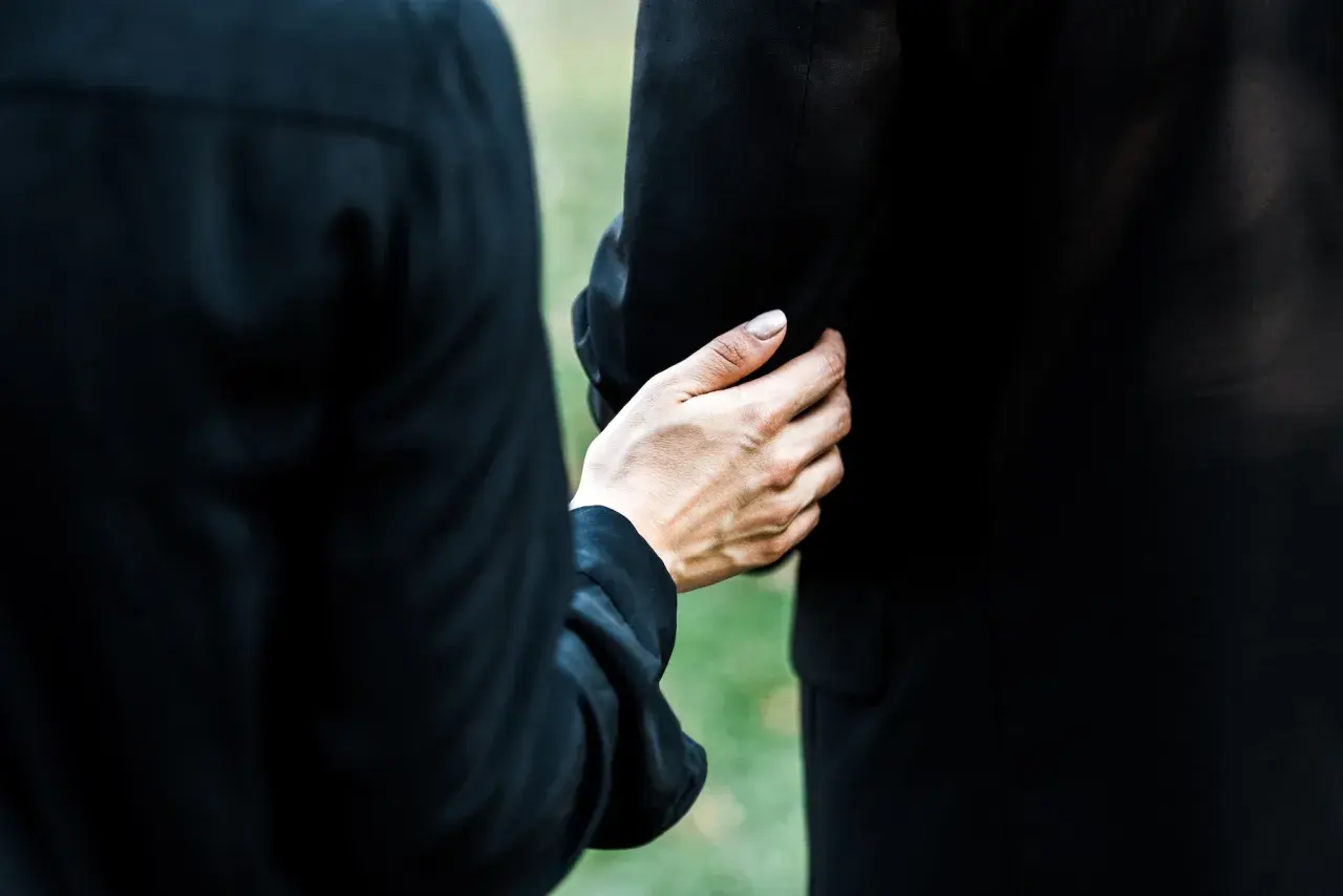 Dłoń obejmuje ramię, wspierając w trudnym momencie. To obraz żałoby i wspólnego przeżywania straty podczas orszaku pogrzebowego.