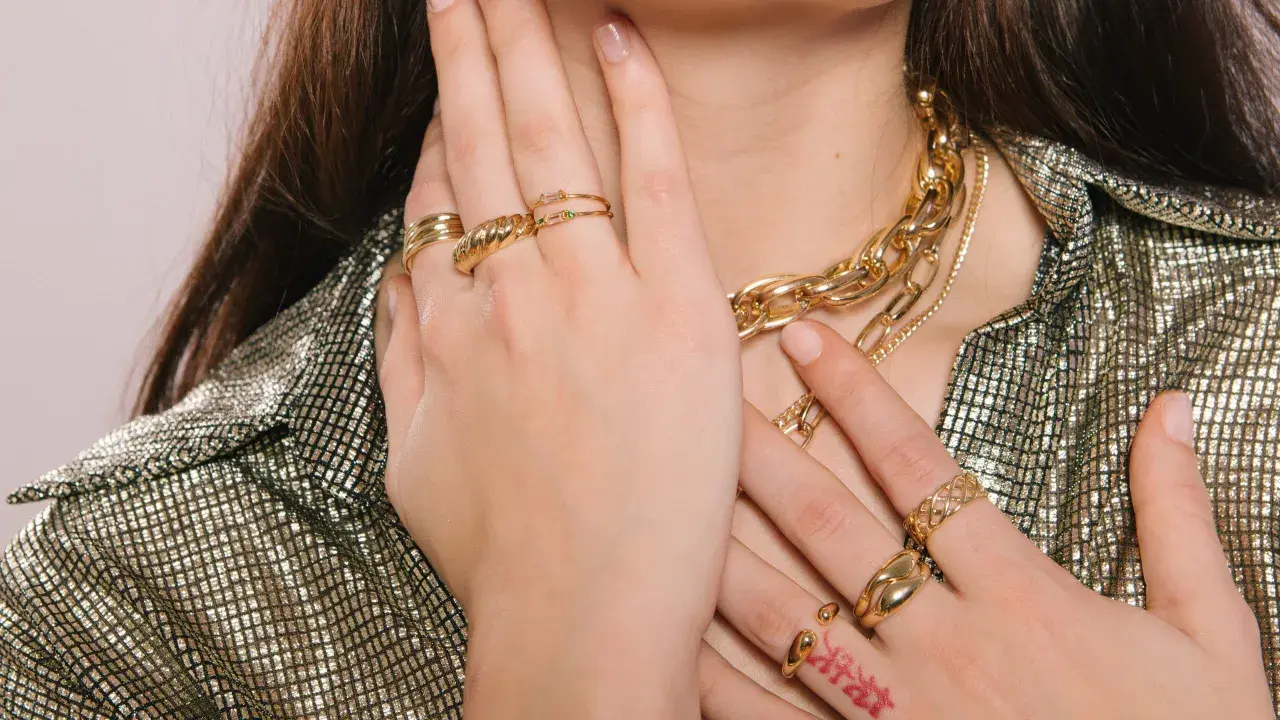 Manos adornadas con varios anillos dorados y un collar grueso, mostrando cómo llevar anillos de manera elegante.
