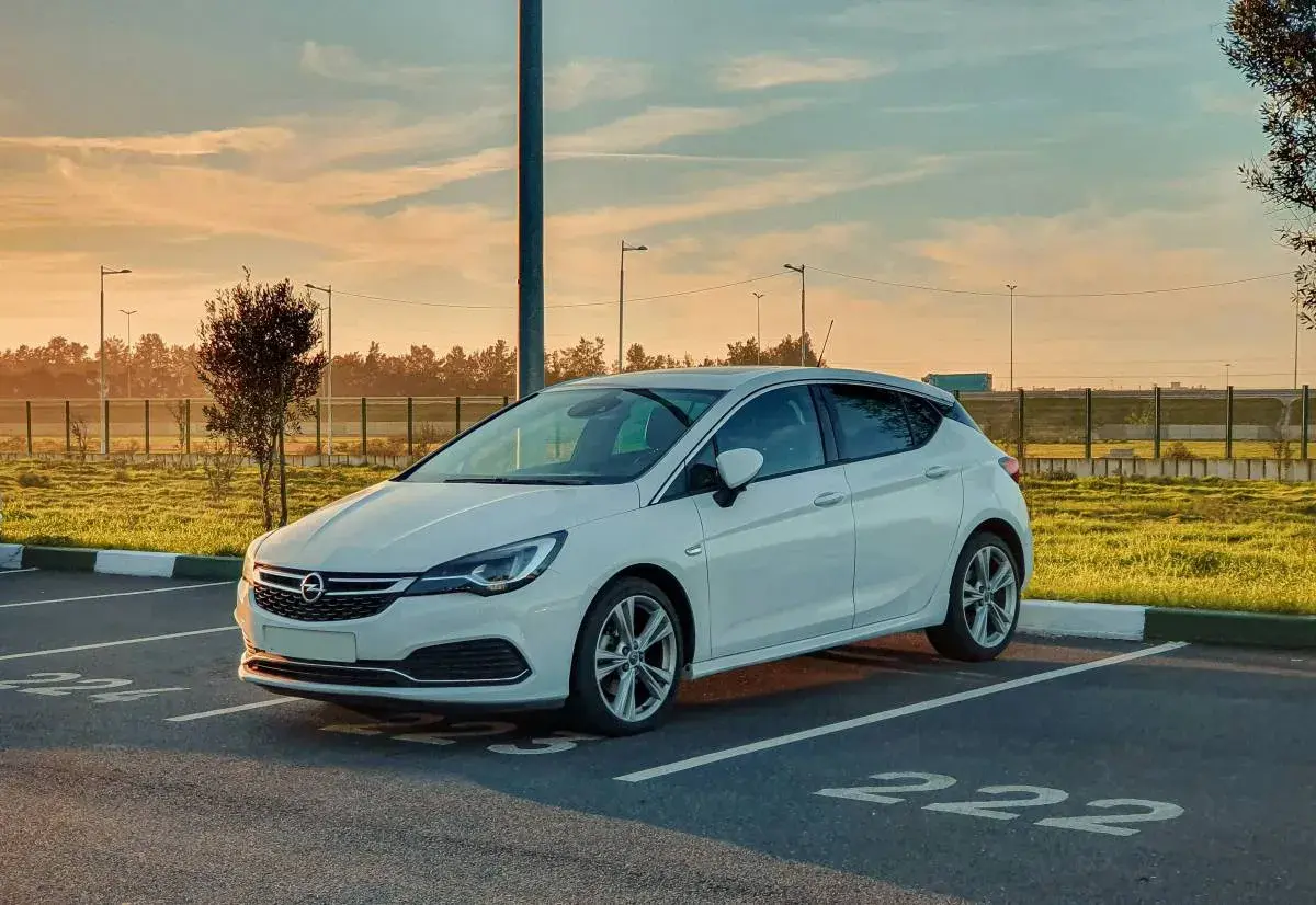 Biała Opel Astra K zaparkowana na parkingu. Słońce zachodzi, tworząc ciepłą atmosferę. Czas na opel astra k wymiana akumulatora.