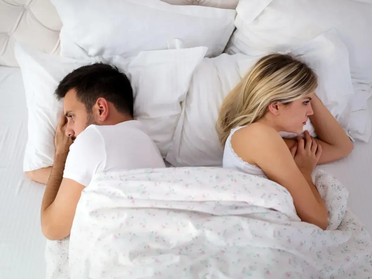 Coppia a letto, distanti. Lei, con lo sguardo perso, sembra non avere voglia di fare l'amore. Lui, pensieroso, si volta dall'altra parte.