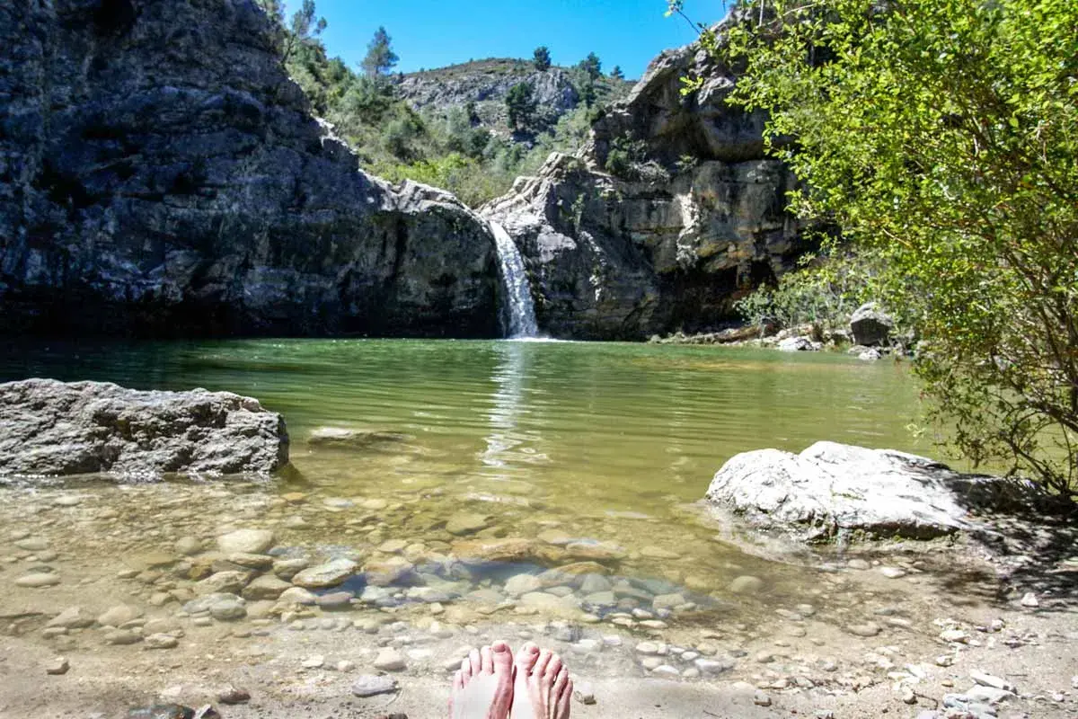 Pies descalzos en la orilla del **barranco de la Encantada**, con una cascada cayendo en una poza de agua verde rodeada de rocas y vegetación.