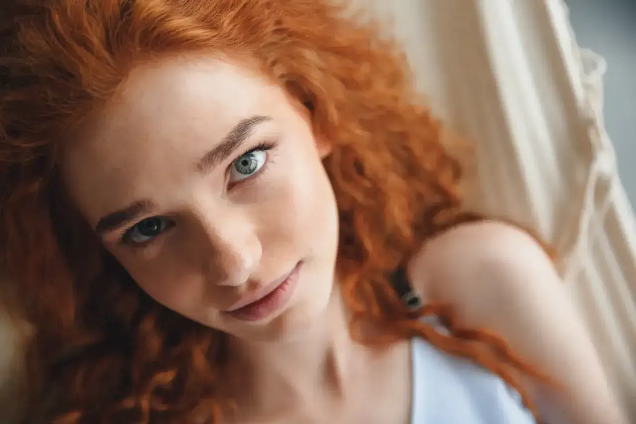 Primer plano de una joven con cabello pelirrojo natural y ojos azules, recostada en una hamaca.