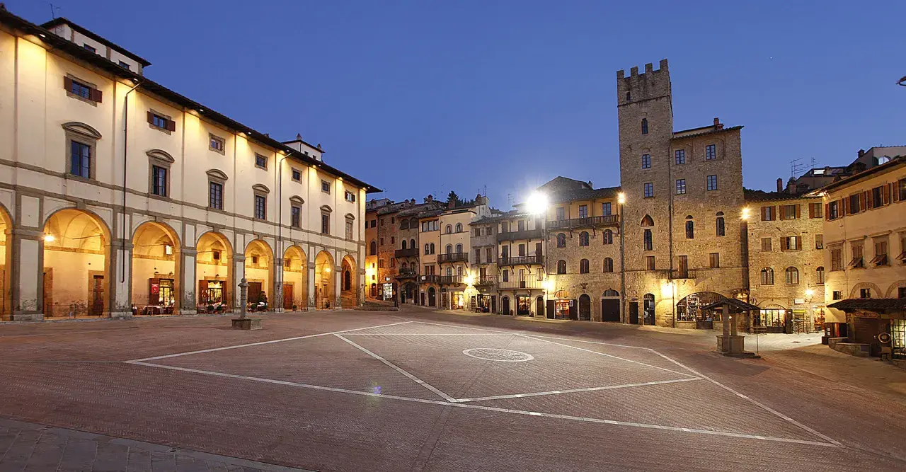 Piazza illuminata ad Arezzo, con portici, edifici storici e una torre. Sembra proprio che la vita è bella ad Arezzo.