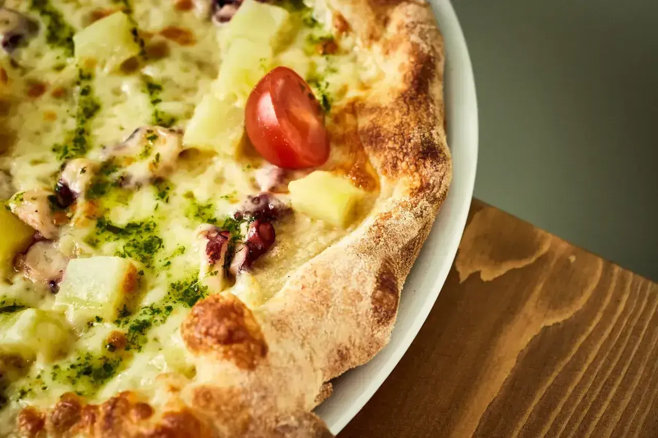 Pyszna pizza z kawałkami ananasa, ośmiornicy i pomidorkiem, obficie posypana zielonym pesto i roztopioną **mozzarellą w bloku**.