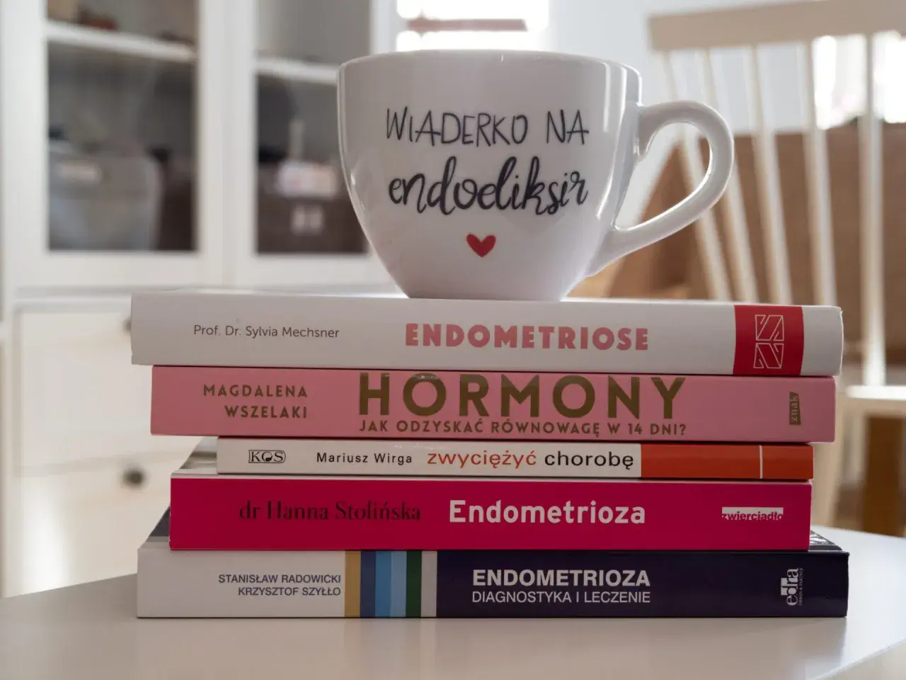 Stos książek o endometriozie, w tym "Endometrioza: diagnostyka i leczenie", z kubkiem "Wiaderko na endoeliksir".