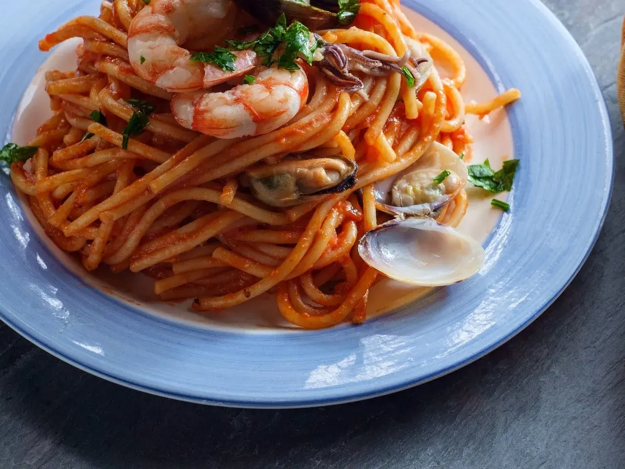 Espaguetis a la marinera con gambas, mejillones y almejas en una salsa roja, servidos en un plato azul.