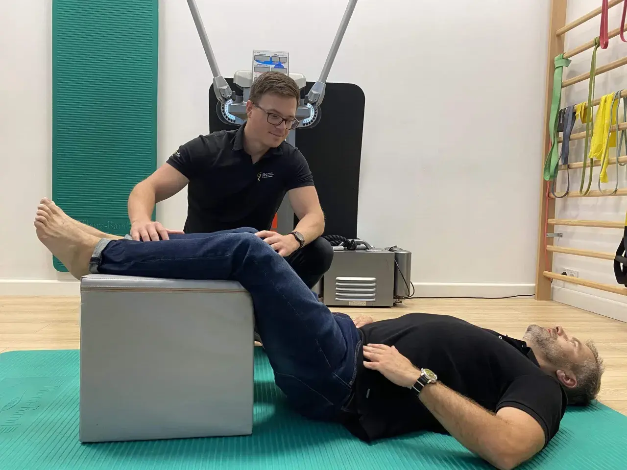 Fizjoterapeuta pomaga pacjentowi w ćwiczeniach na rwę kulszową. Pacjent leży na macie, z nogą opartą na podwyższeniu, a terapeuta wykonuje palpacje.