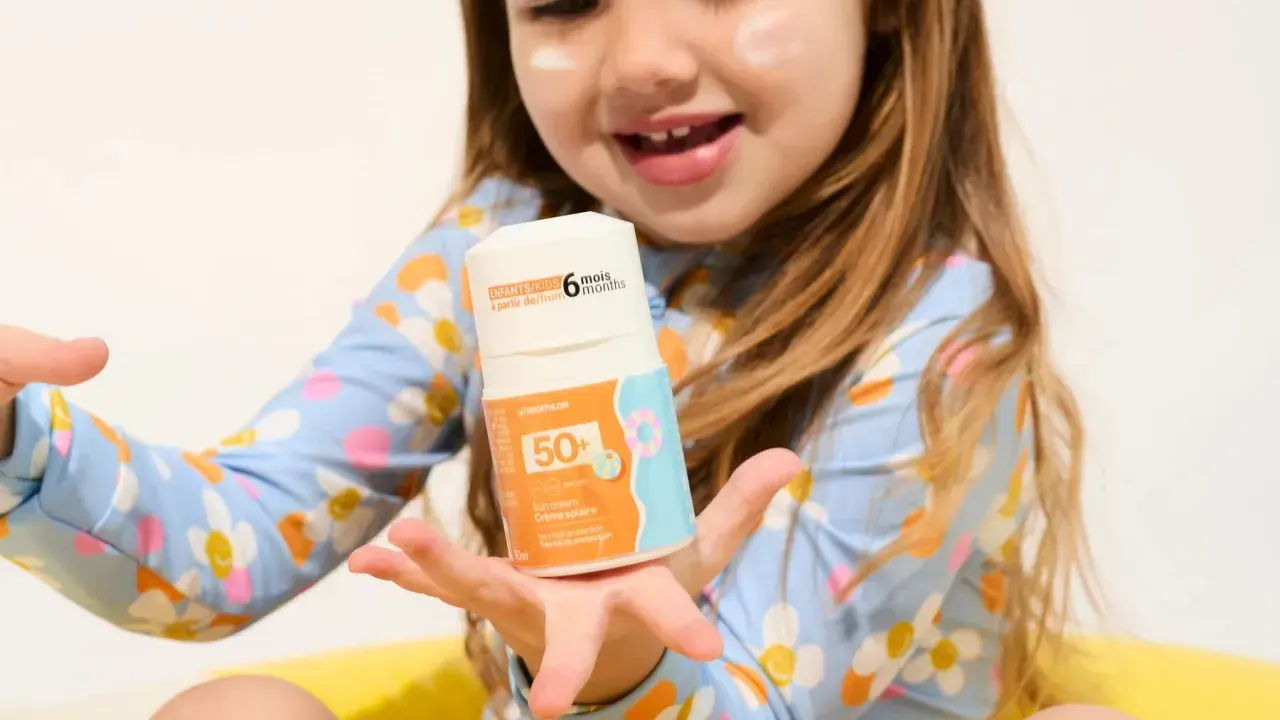 Un enfant tient un flacon de crème solaire UVA UVB 50+. La crème est conçue pour les enfants dès 6 mois.