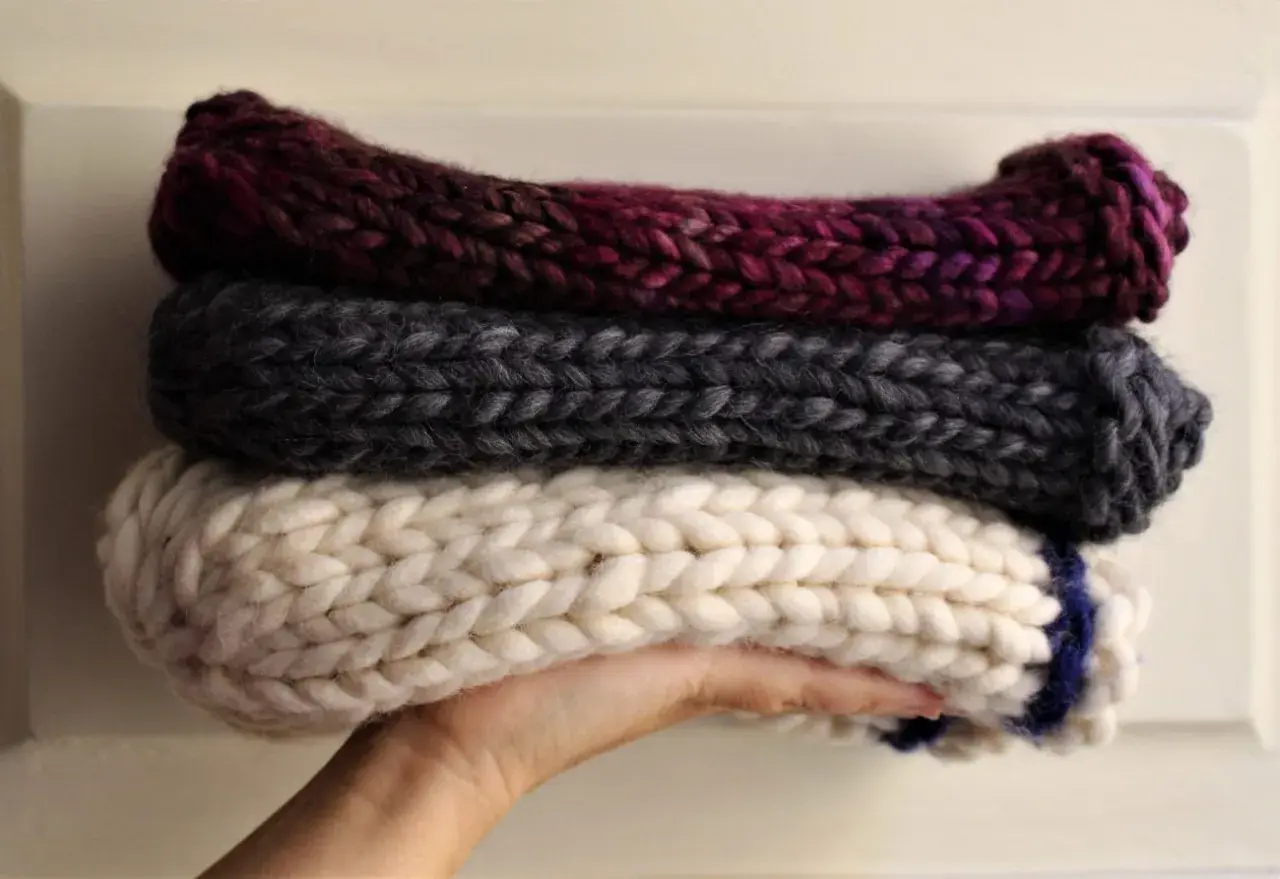 Tre berretti in lana ai ferri, uno bordeaux, uno grigio e uno bianco con striscia blu, impilati su una mano.