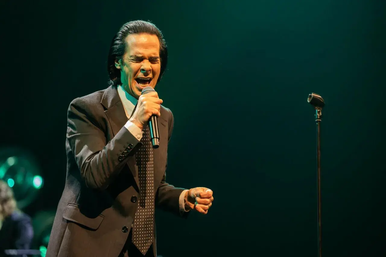 Nick Cave z pasją wykonuje swoje utwory, śpiewając do mikrofonu w eleganckim garniturze.