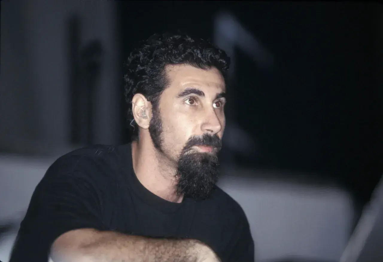 Serj Tankian, wokalista System of a Down, z charakterystyczną brodą i słuchawką w uchu, gotowy do wykonania kolejnych energetycznych utworów.