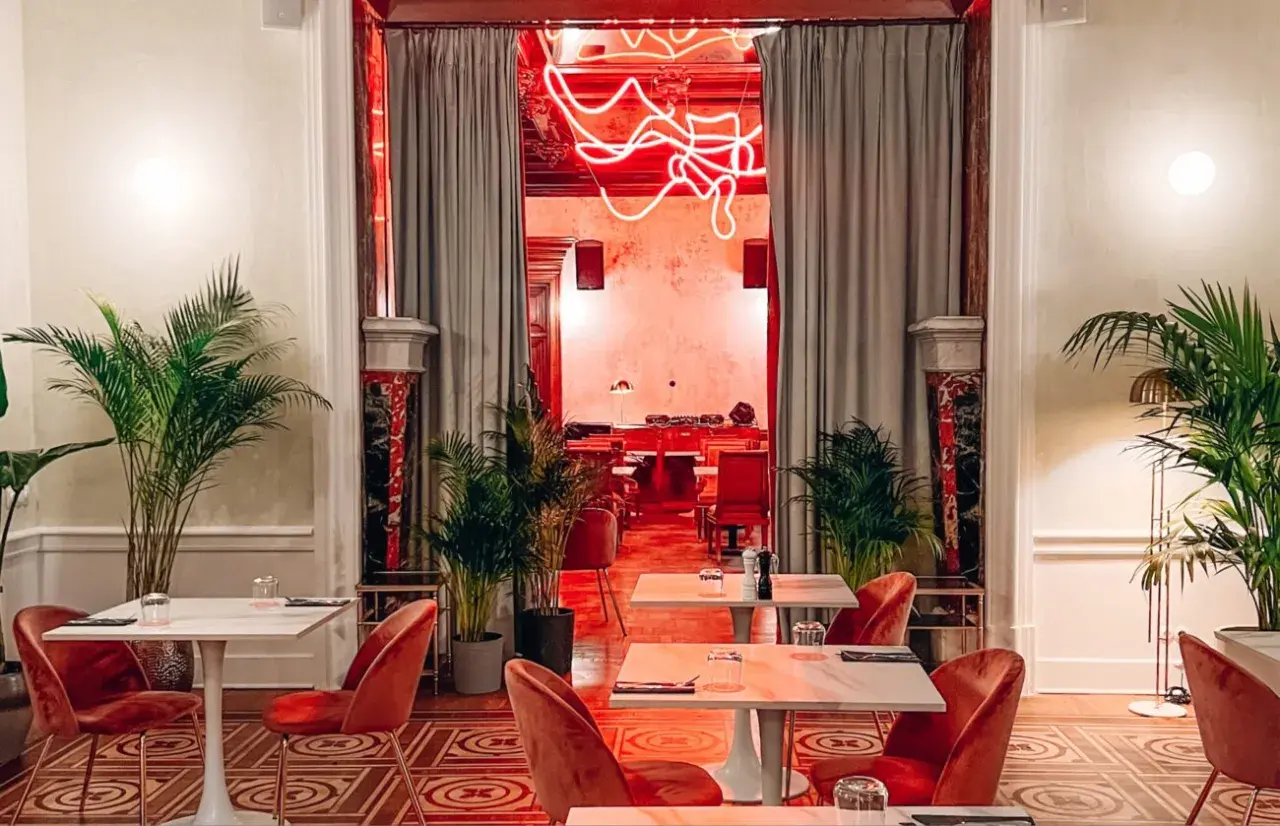 Eleganckie wnętrze z czerwonymi aksamitnymi krzesłami i marmurowymi stolikami. Neonowy napis w głębi sugeruje, że to najdroższa restauracja w Warszawie.