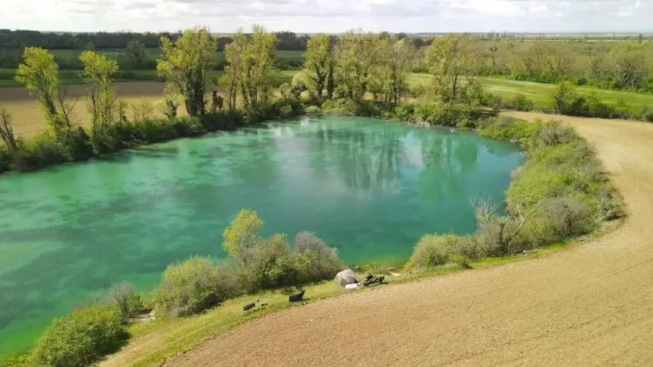 Idyllischer Paylake in Frankreich mit türkisfarbenem Wasser, umgeben von grüner Vegetation und Feldern. Angler haben ihr Lager aufgeschlagen.