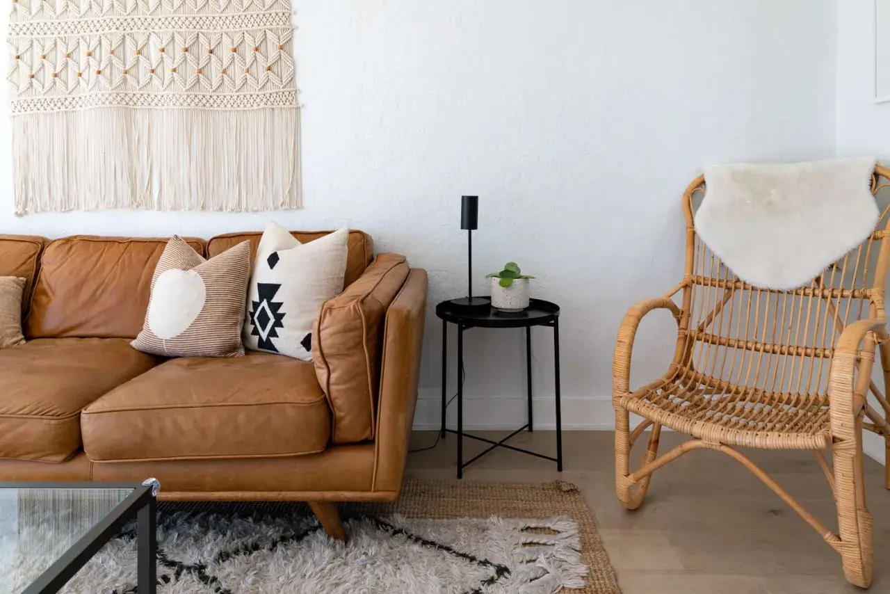 A cozy living room with a brown leather sofa, patterned pillows, a macrame wall hanging, and a rattan chair, embodying a relaxed bohemian furniture style.