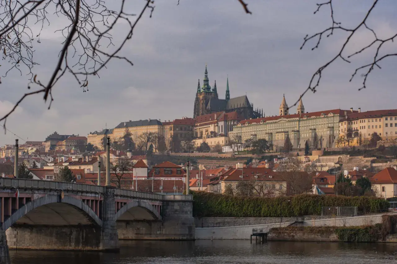 Praga nietypowe atrakcje: widok na Hradczany i Most Karola przez Wełtawę, z gałęziami drzew w pierwszym planie.