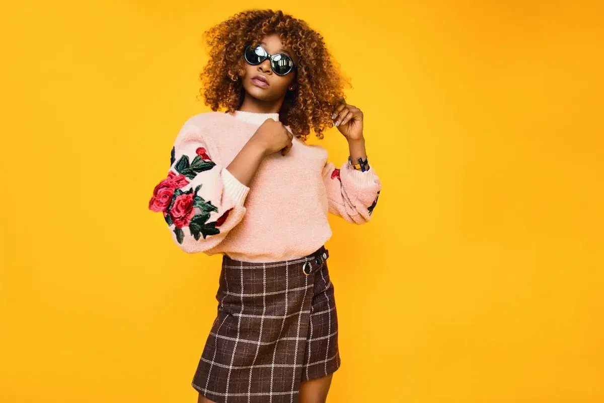 Mujer con cabello rizado y gafas de sol posa con un suéter rosa con rosas bordadas y una falda a cuadros. Su estilo es un armario minimalista moderno.