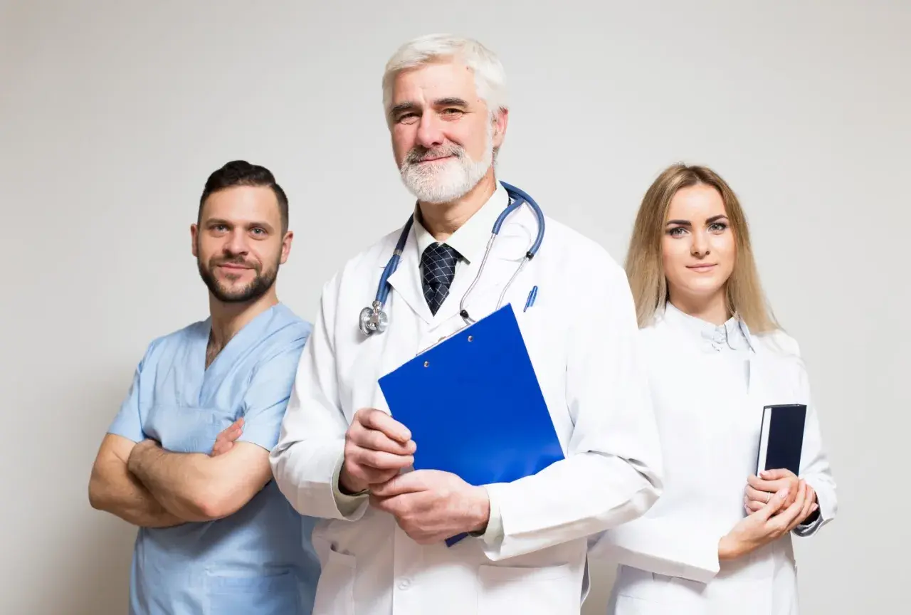 Tre medici sorridono, uno con camice azzurro, uno con camice bianco e stetoscopio, e una donna con camice bianco. Gestiscono la fatturazione medici con professionalità.