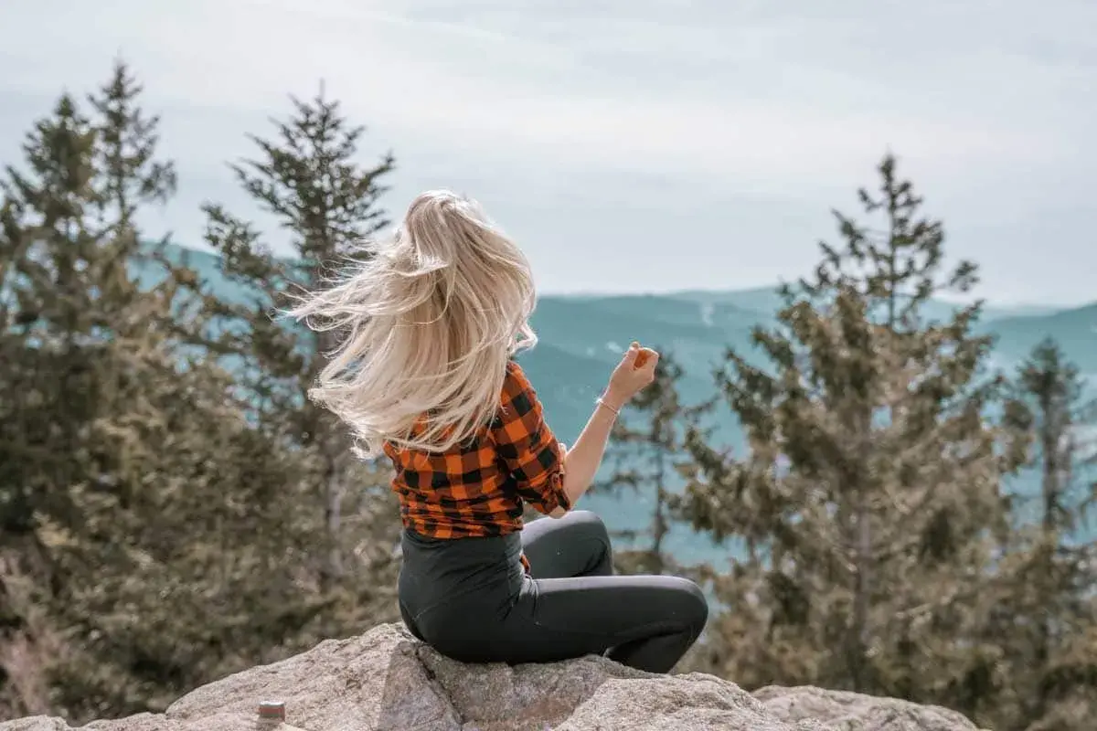 Frau mit wehendem blonden Haar sitzt auf einem Felsen im bayerischen Wald und genießt die Aussicht.
