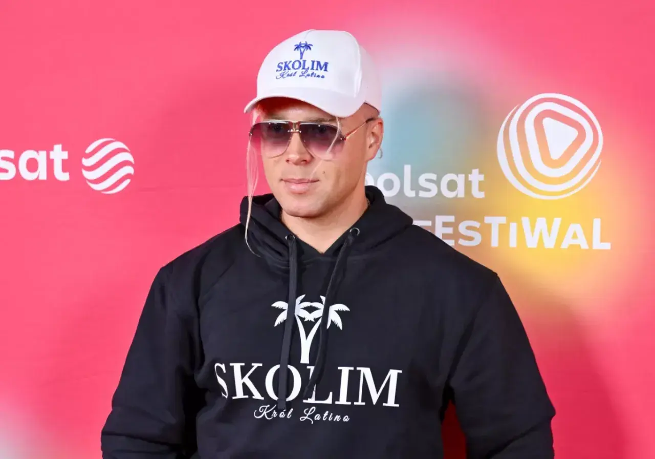 Skolim, Król Latino, w czarnej bluzie z kapturem i białej czapce, emanuje barwami szczęścia na tle festiwalowej ścianki.