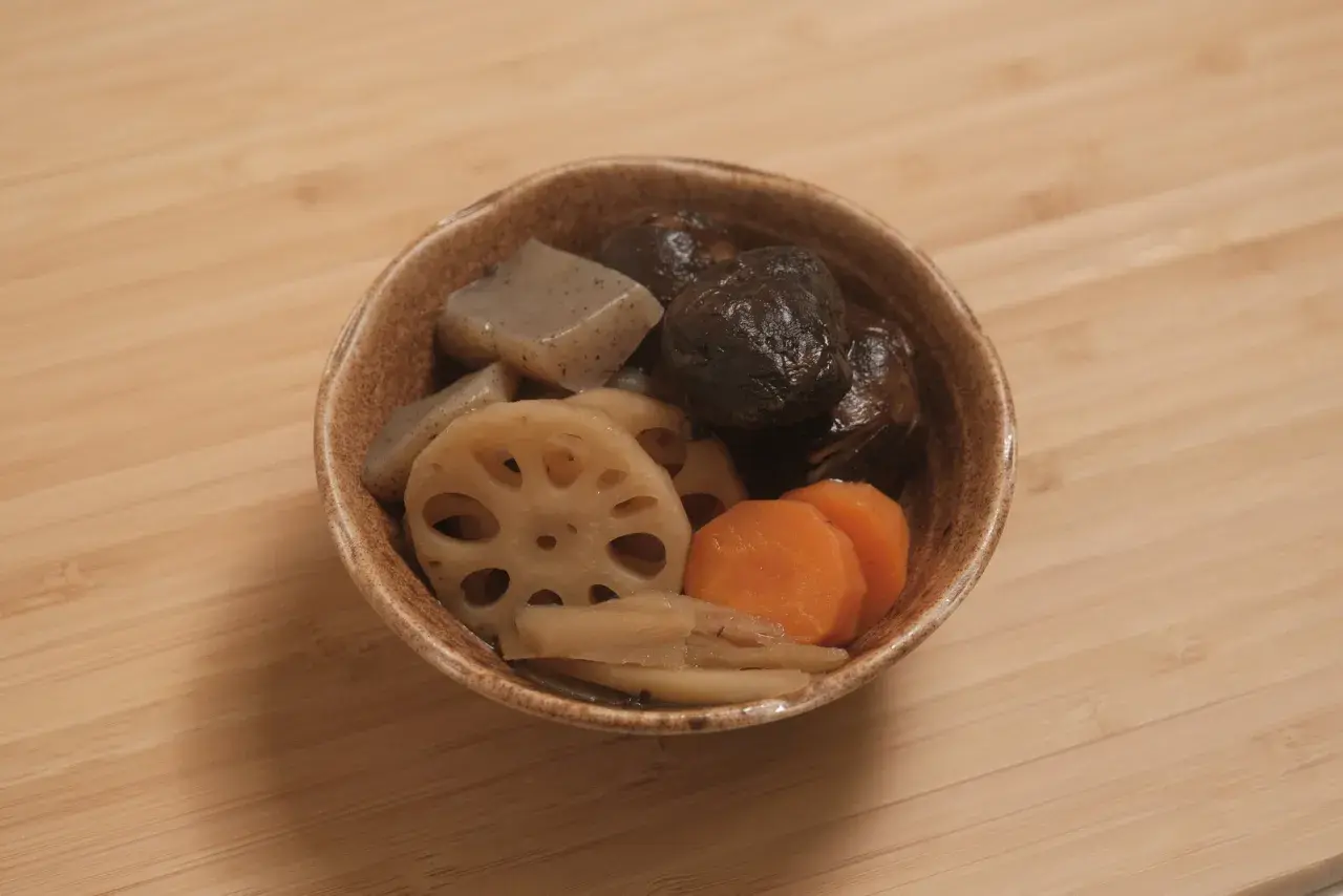 A bowl of simmered vegetables, including shiitake mushrooms, lotus root, carrots, and konjac, likely part of a mushroom dashi broth.