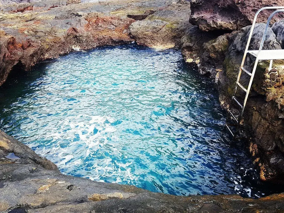 Un charco de los abrigos natural, con aguas cristalinas y una escalera metálica para acceder. Rodeado de rocas volcánicas.