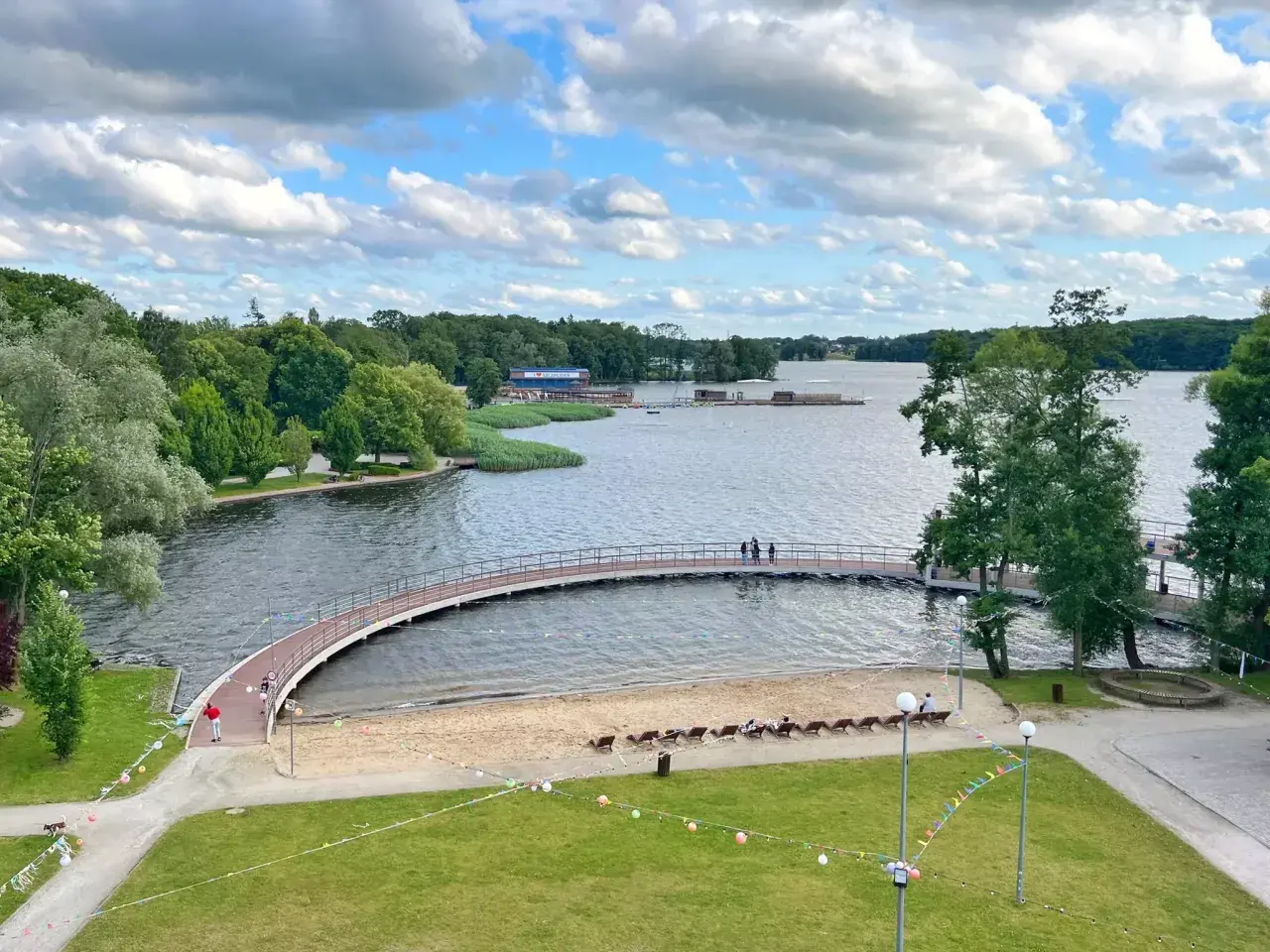 Malownicze jezioro w Szczecinku z łukowatym mostkiem, piaszczystą plażą i zielonymi terenami rekreacyjnymi.