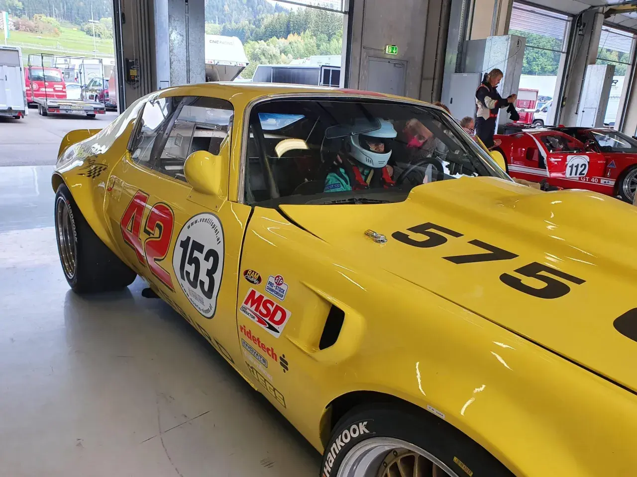 Gul Firebird 69 med nummer 153 och 575 redo för race. En förare i hjälm sitter bakom ratten.