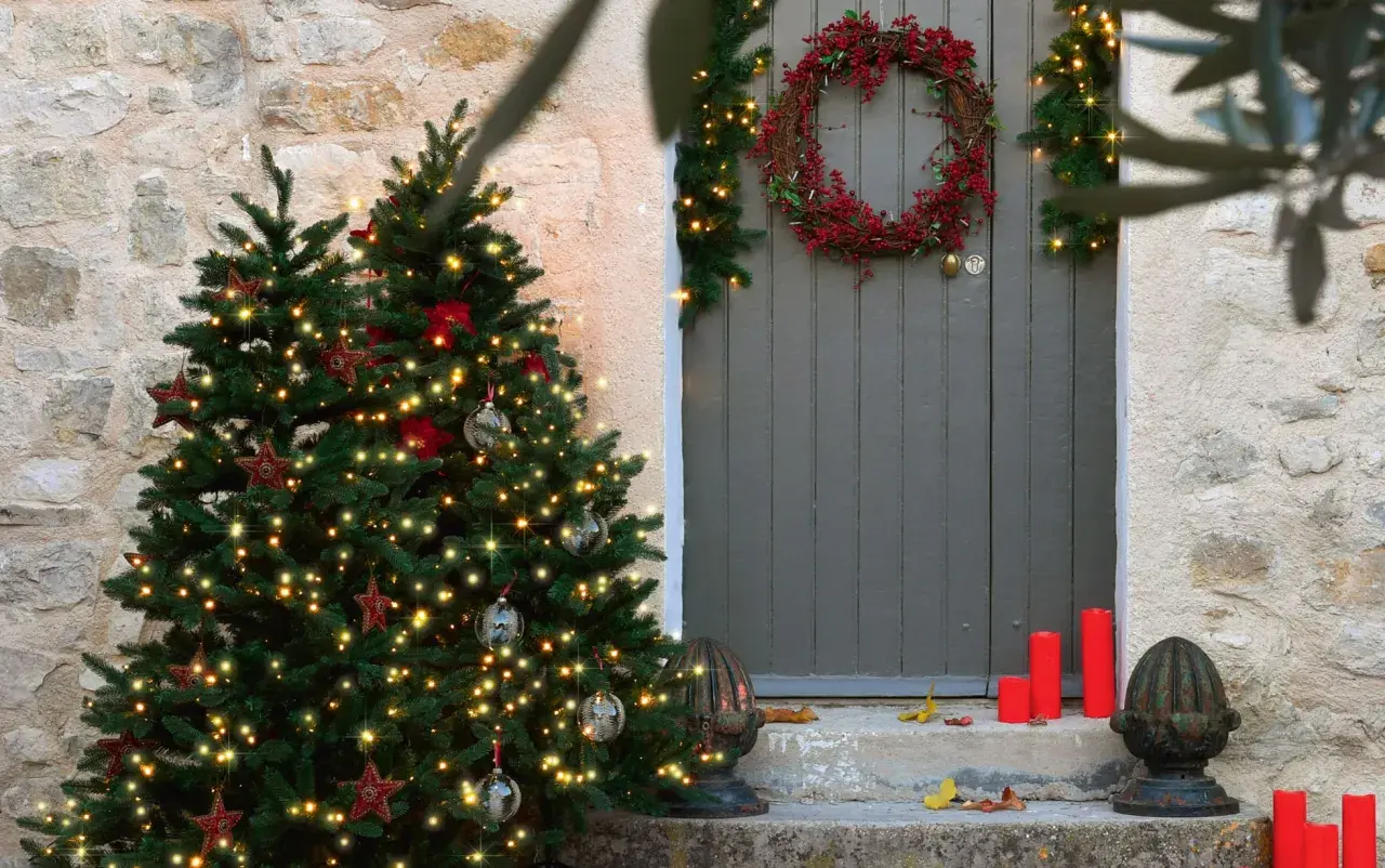 Árbol de Navidad iluminado y una corona de acebo adornan la entrada de una casa de piedra.