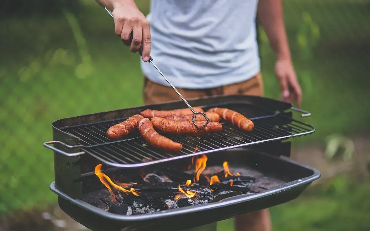 Un homme retourne des saucisses sur un barbecue bois ou charbon. Les flammes dansent sous la grille, promettant un repas savoureux.