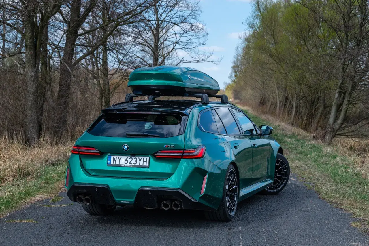 Zielone BMW M5 Touring z boxem dachowym na tle drzew i nieba.