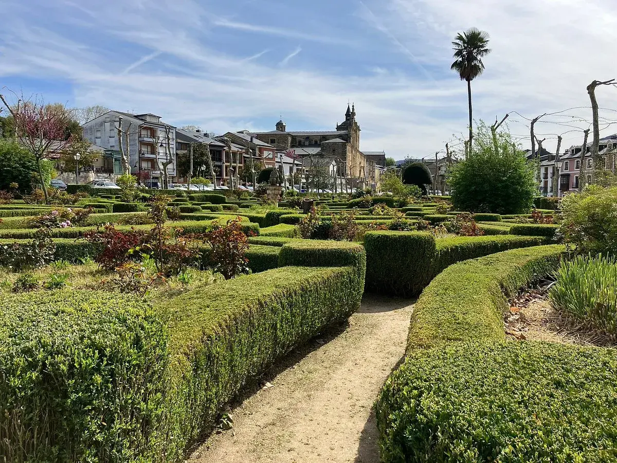 Jardín geométrico con setos cuidados y flores en Villafranca del Bierzo. Al fondo, edificios históricos y una palmera solitaria bajo un cielo azul.