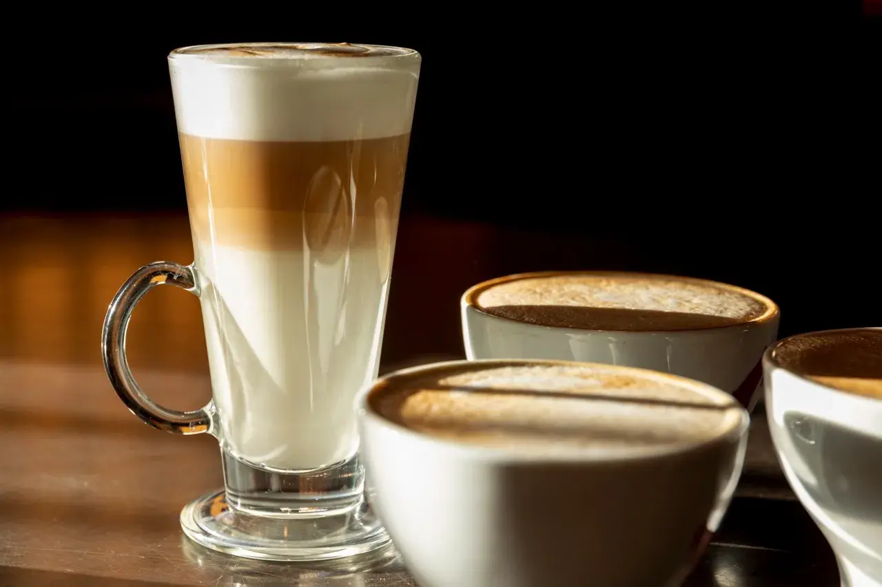 Kawa biała w wysokiej szklance z warstwami mleka i espresso, obok trzy filiżanki kawy z pianką.