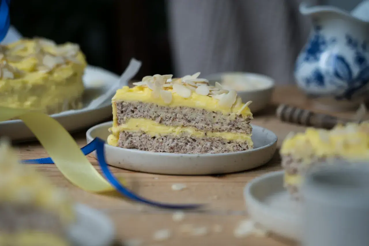 Ein Stück schwedische Mandeltorte mit gelber Creme und Mandelblättchen, dekoriert mit blauen und gelben Bändern.