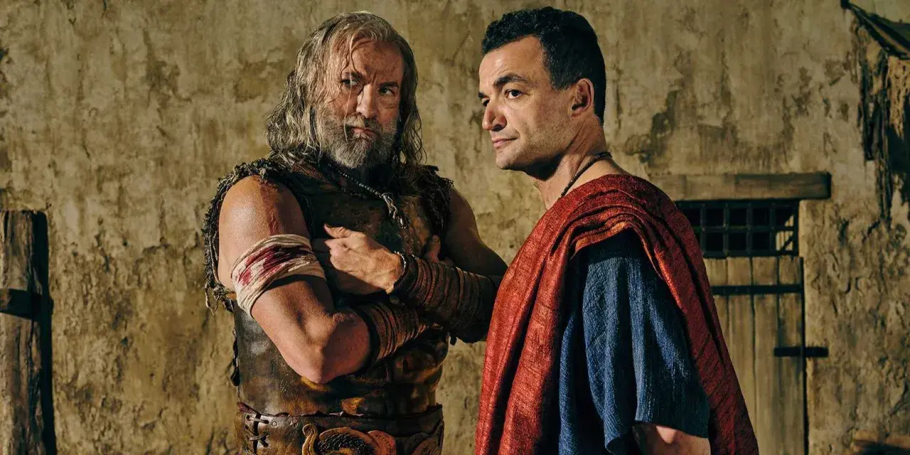 Två män i antika kläder, en med skägg och läderrustning, den andra i röd mantel, står nära varandra. Kanske en scen från Spartacus: House of Ashur.