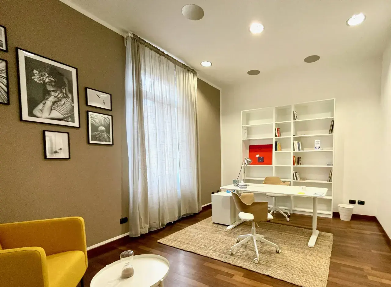 Arredamento studio psicologo: scrivania bianca, libreria, sedie, poltrona gialla, quadri e tenda.