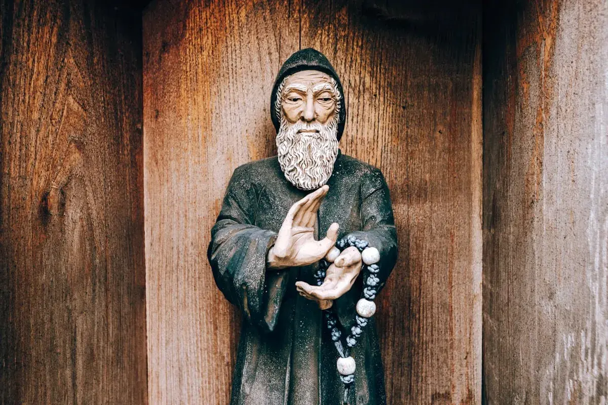 Posąg mnicha z brodą i szarbel uzdrowienia w dłoniach, na tle drewnianej ściany.