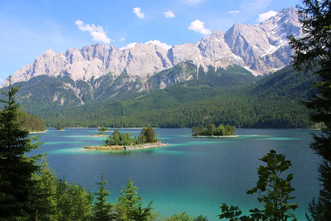 Turquoise lake with islands dotted with trees, framed by a dense green forest and majestic, snow-capped mountains. A quintessential view of the Bavarian countryside.
