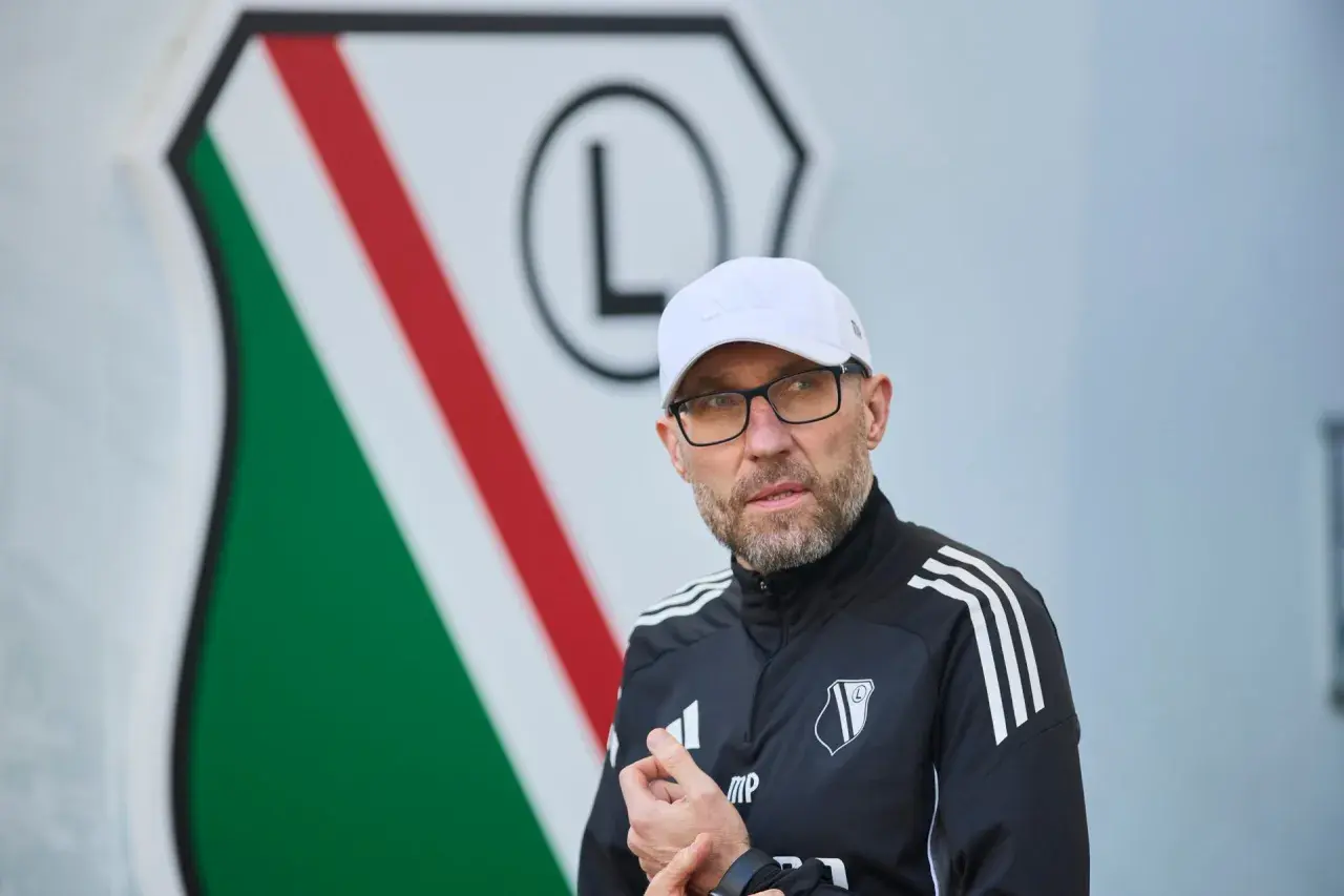 Nowy trener Legii w białej czapce i okularach, na tle klubowego herbu.