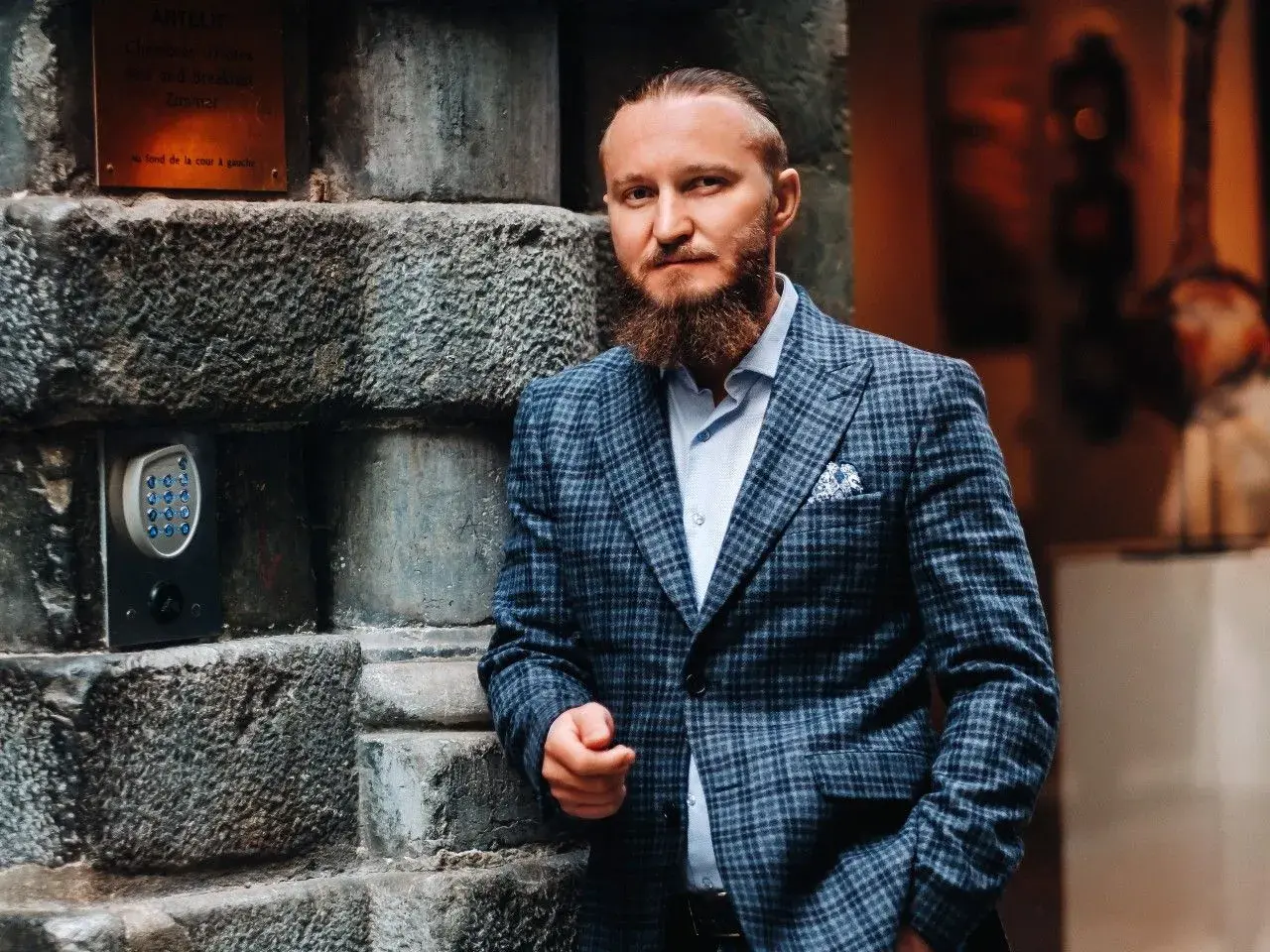 Elegancki mężczyzna w garniturze w kratę, z brodą, oparty o kamienną ścianę. Idealny przykład, jaki krawat do garnituru w kratę wybrać.