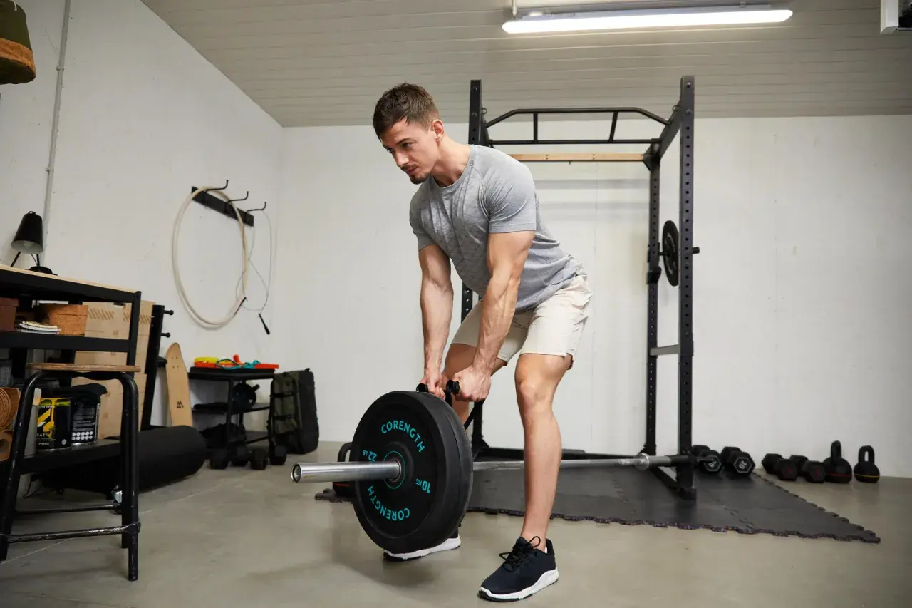 Homme musclé en buste penché, soulevant une barre chargée de poids dans une salle de sport à domicile.