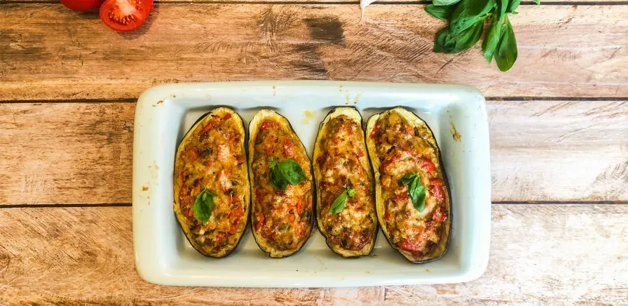 Quatre moitiés d'aubergines farcies, gratinées et garnies de basilic, dans un plat blanc sur fond de bois.