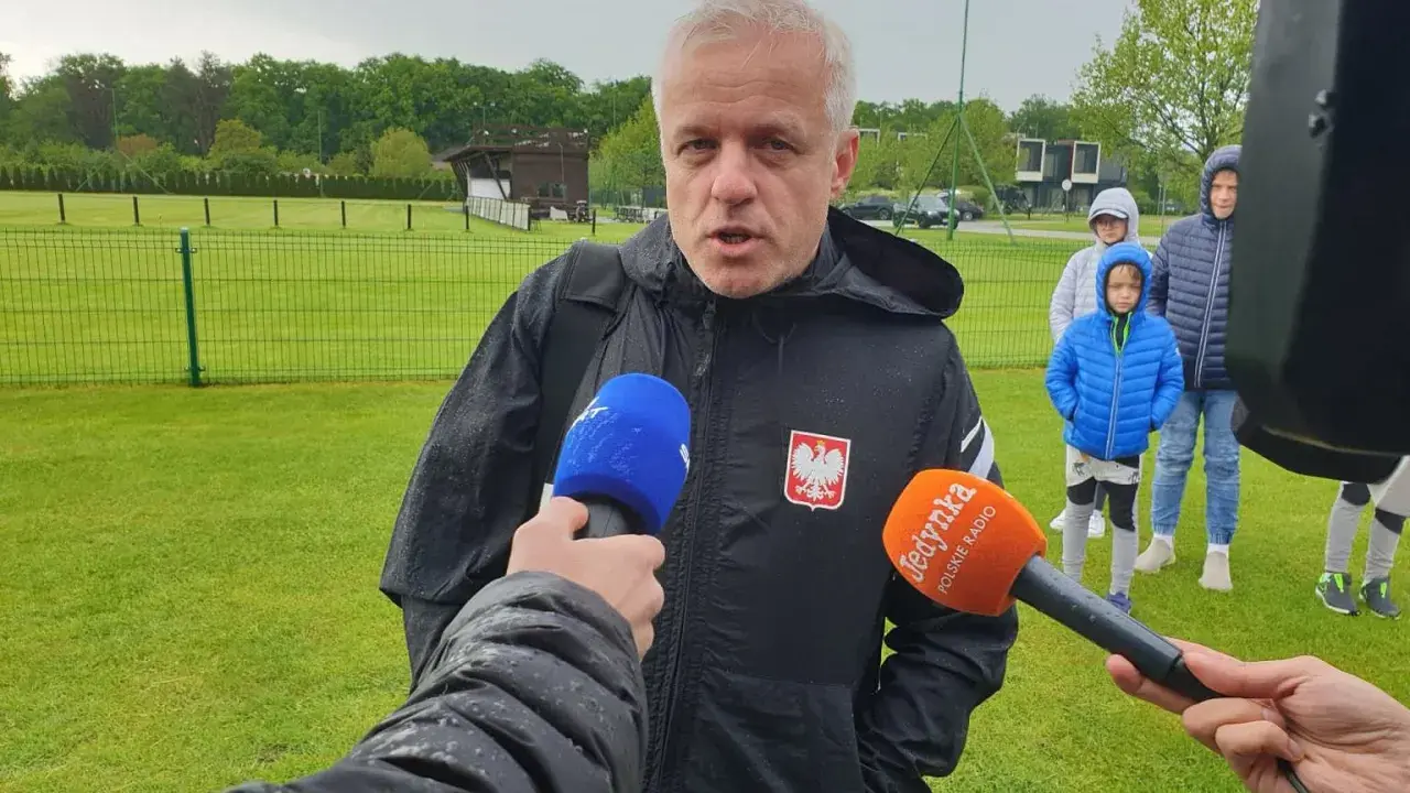 Jacek Jaroszewski, w czarnej kurtce z orłem, udziela wywiadu na boisku. W tle młodzi piłkarze.