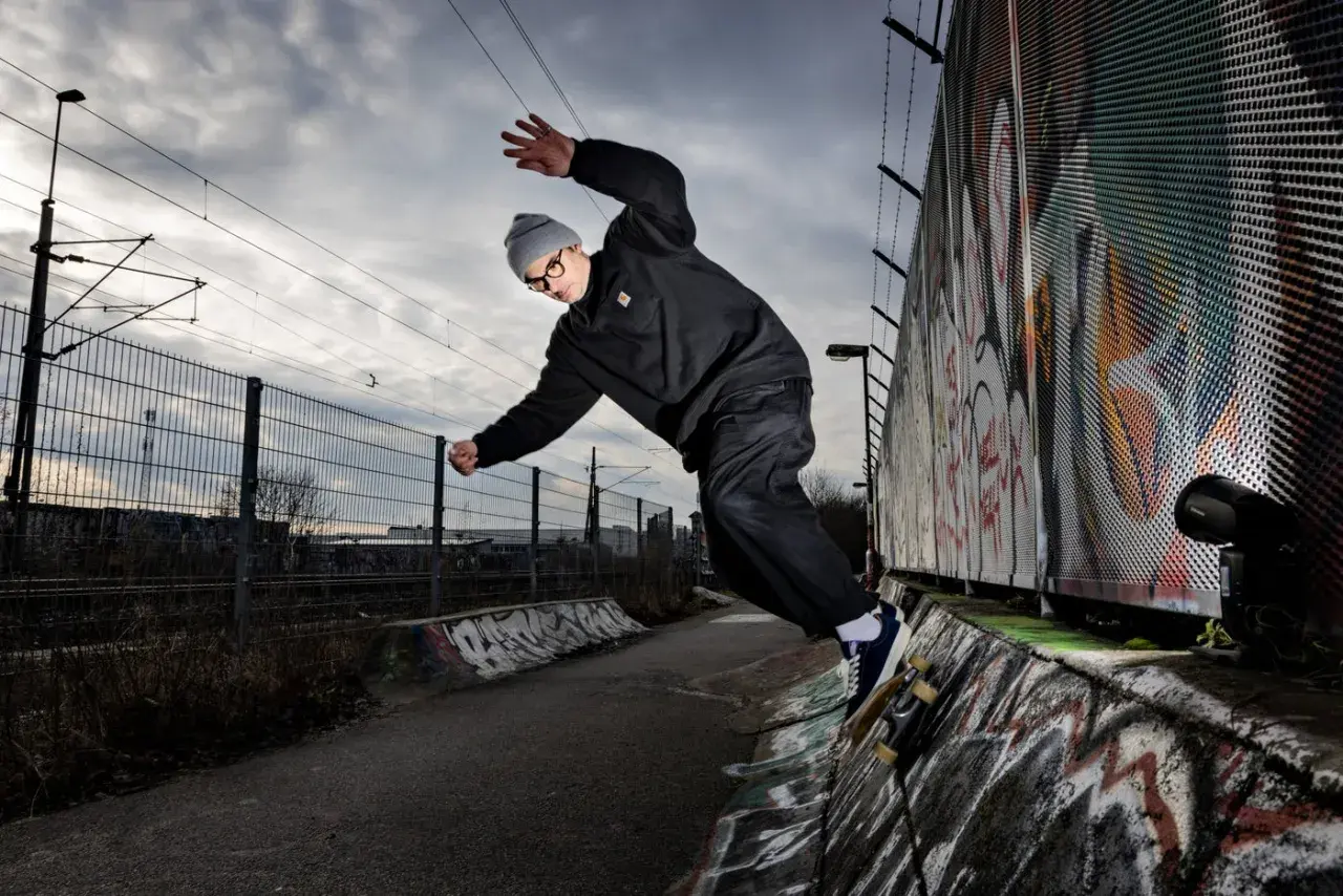 Martin Ander gör ett trick på skateboard i en betongpark med graffitiväggar och järnvägsspår i bakgrunden.