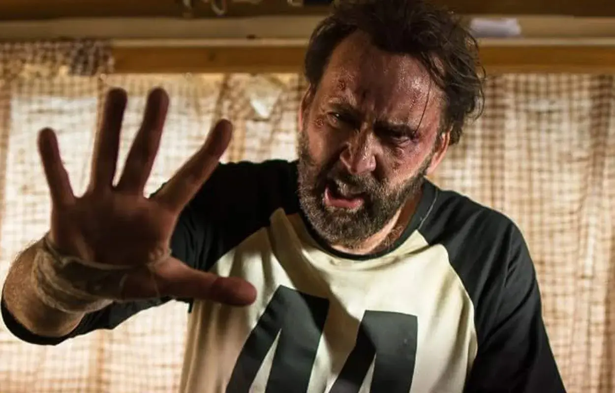 Nicolas Cage med blåmärken och svettig panna, höjer handen i en gest av stopp. En scen från en av hans filmer.