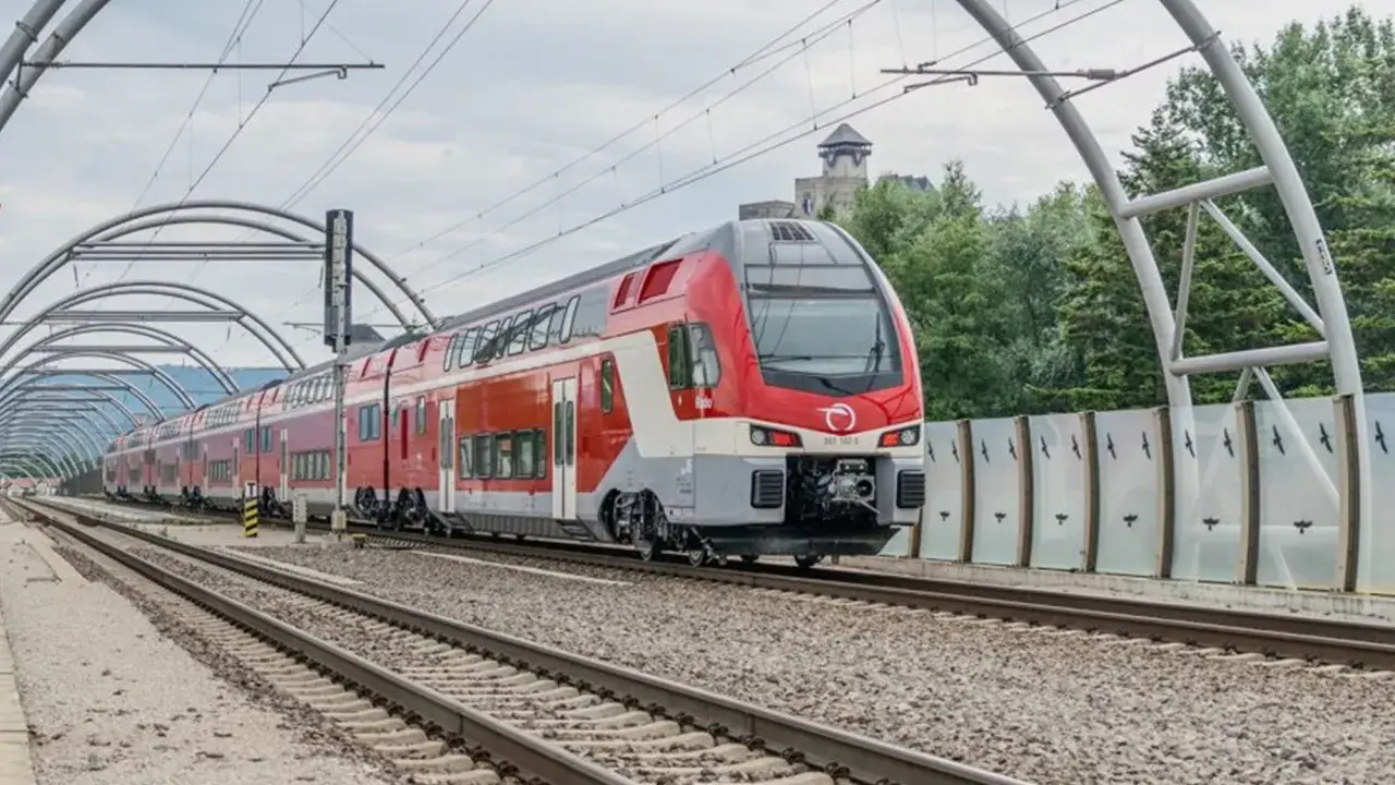 Moderner Doppelstockzug der **Slovak Railways** fährt unter einer Bogenkonstruktion auf Gleisen. Im Hintergrund eine Burg.
