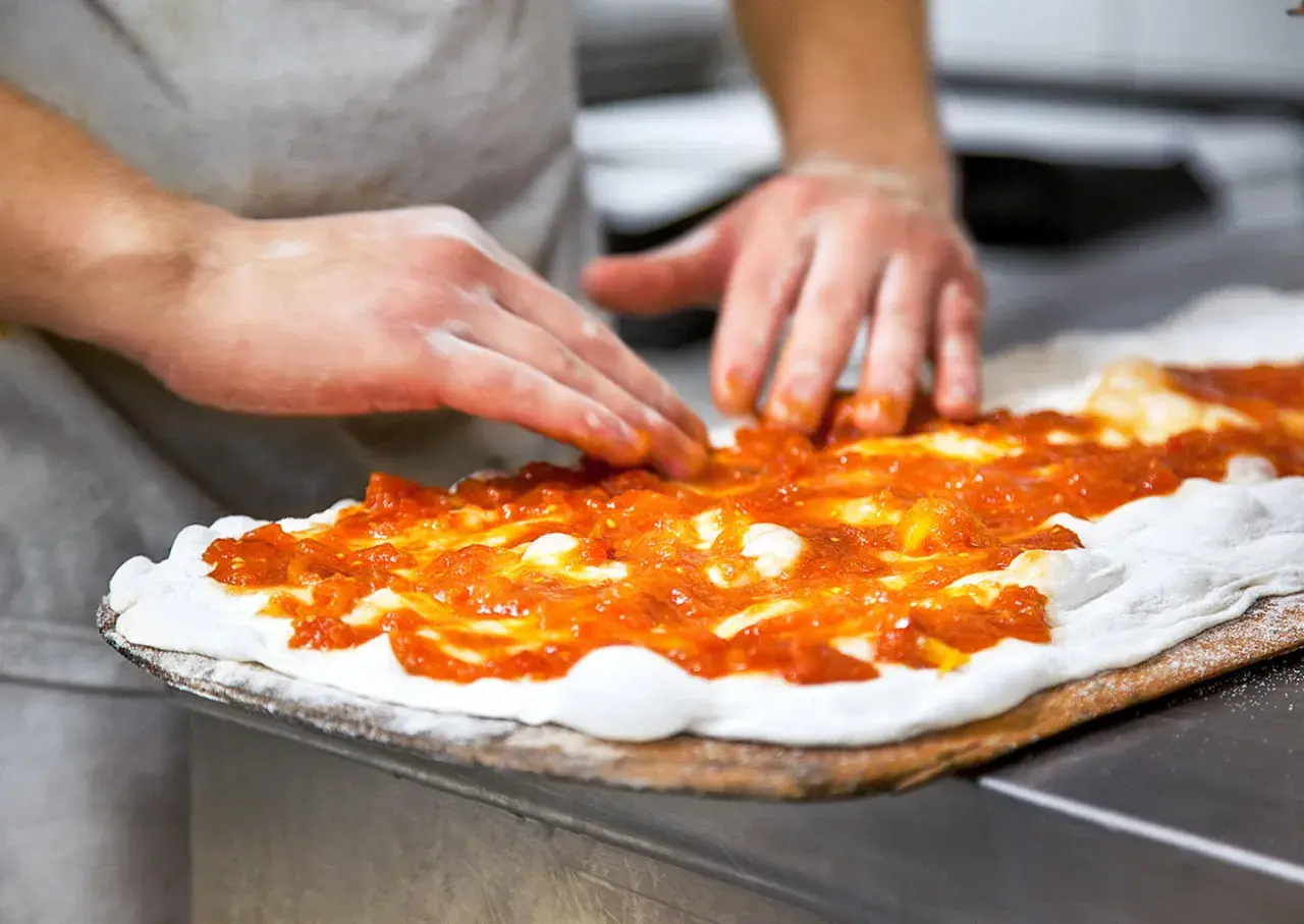Händer sprider tomatsås på pizzadeg. En del av en matlagningskurs i att baka pizza.