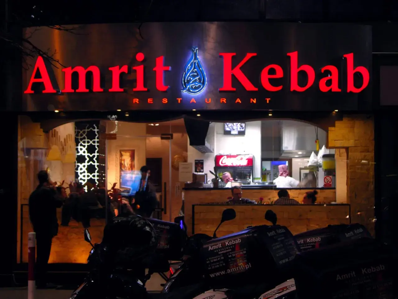 Jasny neon "Amrit Kebab" nad wejściem do restauracji. W tle widać wnętrze z pracującymi kucharzami i lodówką Coca-Coli. Na pierwszym planie stoją skutery z pudełkami na dowóz.