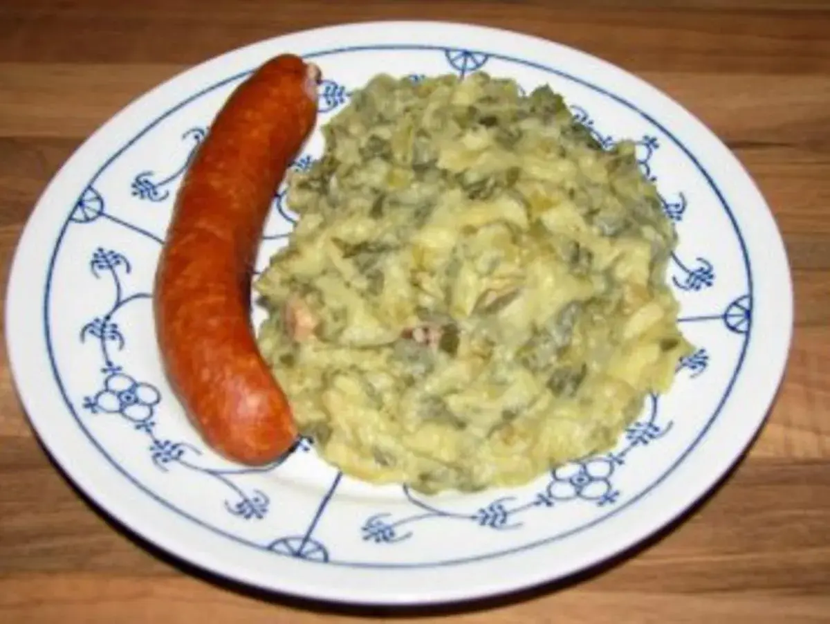 Omas Stielmus Rezept: herzhafte Grünkohl-Kartoffel-Masse mit einer deftigen Bratwurst auf einem Teller mit blauem Muster.