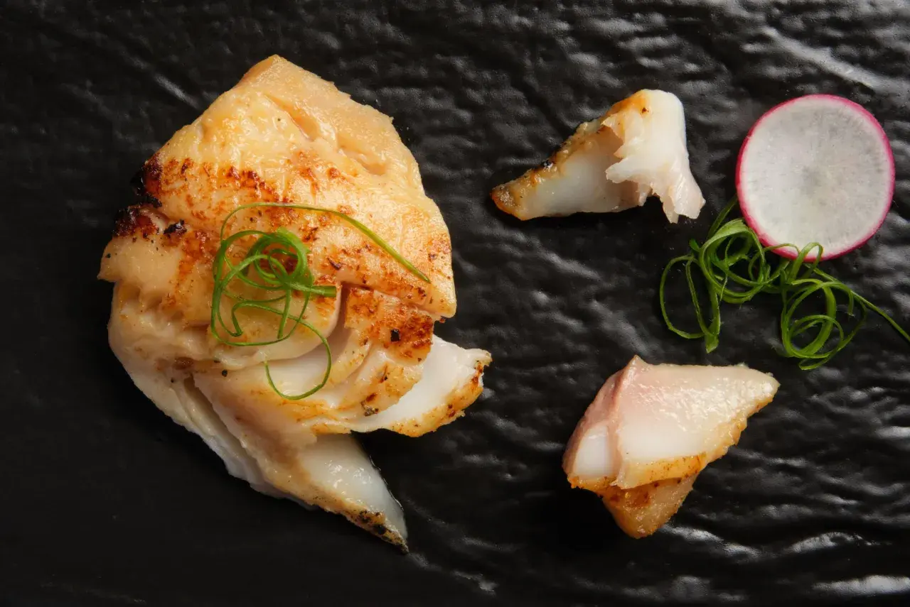 A beautifully grilled saikyo yaki fish fillet, garnished with green onions and a radish slice, rests on a black plate.