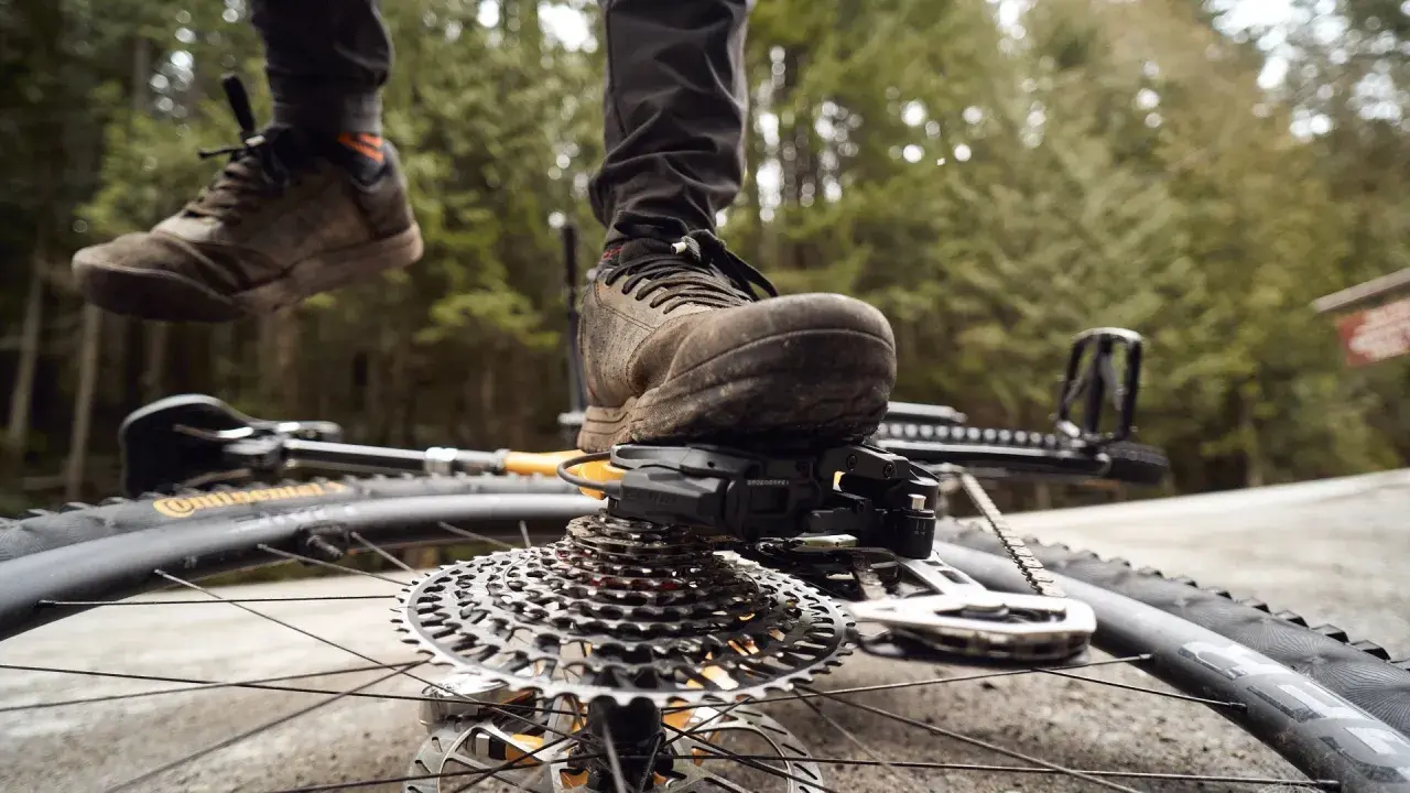 A mountain biker's muddy shoe rests on the SRAM T-Type derailleur and cassette of a bike lying on its side.